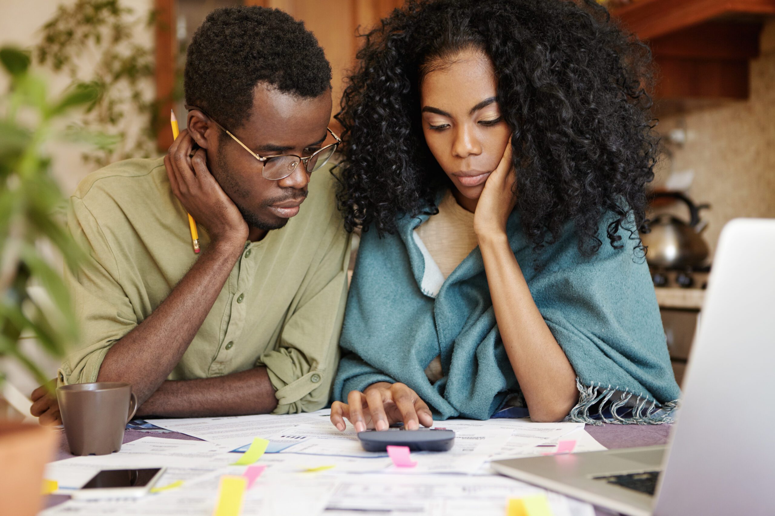 Stressed young African couple can’t stand tension of financial crisis, looking unhappy and frustrated, sitting at kitchen table with calculator, trying to save some money by cutting domestic expenses